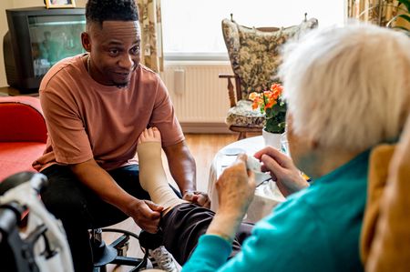 Een man kijkt vriendelijk naar de vrouw terwijl hij haar kous aantrekt. Ze zitten in een gezellige woonkamer met meubels met bloemenprints en een plantje op tafel.