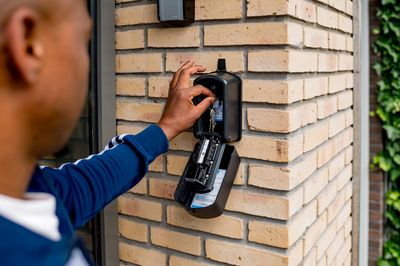 Een man met blauwe trainingsjas staat bij een bakstenen muur en sluit een sleutelkastje dat aan de muur hangt.