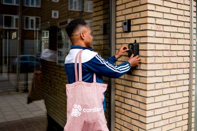 Een man met blauwe trainingsjas en lichtrode, canvas Cordaan tas staat bij een bakstenen muur en sluit een sleutelkastje dat aan de muur hangt.