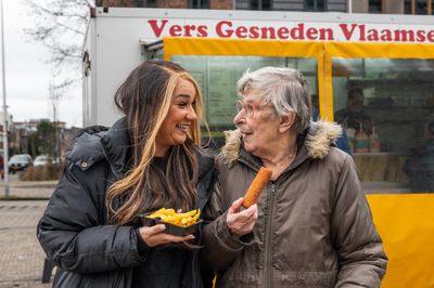 Medewerker met friet en cliënt met kroket staan voor frietkraam en lachen naar elkaar.