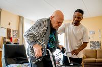Een jonge man met donker haar en wit shirt helpt een oudere, kale man met grijs geprint vest met het lopen met zijn rollator. Ze staan in een woonkamer met lichtoranje muren.