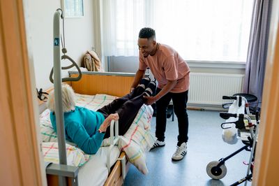 Een jonge man lacht vriendelijk terwijl hij een oudere dame haar bed in helpt. Hij tilt haar benen in het bed en de vrouw houdt zich vast aan een steun aan het bed.
