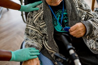 Een man met een grijs vest met print zit in een zwarte leren stoel en houdt met een hand zijn alarmketting vast. De zorgmedewerker draagt blauwe latex handschoenen en heeft zijn handen op de arm van de man.