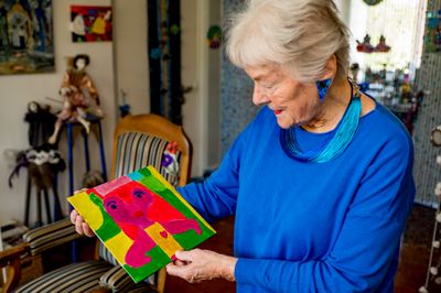 Oudere dame met blauwe trui en blauwe accessoires kijkt naar een kleine kleurrijke schilderij van een meisje in haar hand