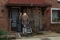 Medewerker en cliënt met rollator lopen vanuit huis naar buiten