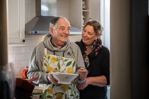 Oudere man met schort en kom in zijn hand en medewerker in de keuken.