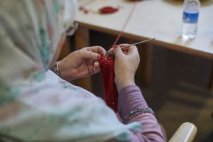 Mevrouw met een hoofddoek en een bril haakt met rood garen.