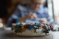 Op de voorgrond een krans met vlinders van kralen. Op de achtergrond iemand die een naald en draad vast heeft.