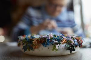 Op de voorgrond een krans met vlinders van kralen. Op de achtergrond iemand die een naald en draad vast heeft.