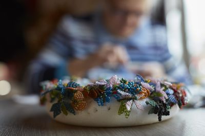 Op de voorgrond een krans met vlinders van kralen. Op de achtergrond iemand die een naald en draad vast heeft.