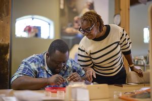 Een vrouw kijkt mee over de schouder van een man die aan tafel iets aan het maken is.