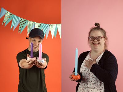 Twee portretfoto's naast elkaar van een jongen en een meisje met kandelaren met kaarsen in hun hand. De achtergrond is kleurrijk met vlaggetjes.