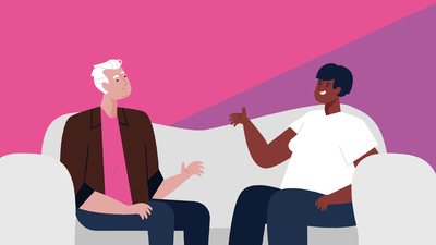 Een kwieke man met wit haar en bruin jasje zit links op een bank. Rechts zit een dame met zwart haar. Ze zijn in gesprek.