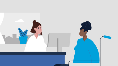 Een dame met blauwe trui in rolstoel zit tegenover een vrouw met bruin haar en een knotje met wit uniform aan. Op haar bureau staat een beeldscherm