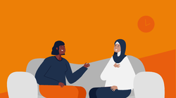 Twee dames zitten op een grijze bank en zijn in gesprek. De een heeft halflang zwart golvend haar, de ander een wit shirt en donkerblauwe hoofddoek