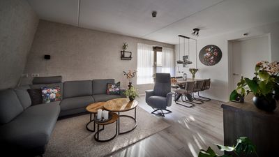 Een moderne woonkamer met een grijze bank, ronde houten tafels, een eethoek en planten.