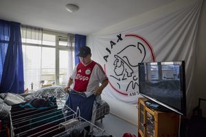 Een man met een voetbalshirt aan hangt de was op in zijn woning, aan de muur hangt een grote voetbalvlag.