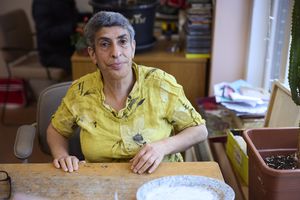 Een vrouw met kort haar en gele blouse zit aan tafel en kijkt in de camera. Op tafel ligt een keramieken geschilderd bord.