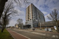 Een hoog, modern appartementencomplex aan een brede straat met fietspaden, omgeven door bomen onder een gedeeltelijk bewolkte hemel.