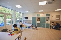 In een kleurrijk klaslokaal met grote kast met blauwe kastdeuren zijn kinderen aan het spelen onder begeleiding van medewerkers.
