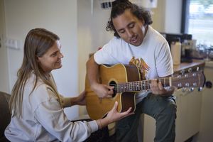 Een man speelt op een gitaar, een vrouw zit tegenover hem en houdt de gitaar vast.
