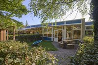 Een gelijkvloers gebouw met grote ramen, omgeven door bomen en een tuin, met een zitgedeelte buiten en een speeltoestel.