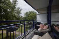 Een breed balkon met zitmeubelen en kussens, het balkon kijkt uit over een park met gras en bomen.