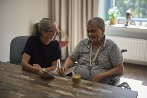 Een bewoner en een begeleider zitten aan tafel en de bewoner kijkt mee op de telefoon van de medewerker.