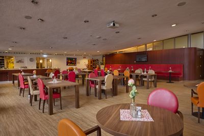 Ruime eetruimte met houten tafels, kleurrijke stoelen en bloemstukken op tafel. Langs de muur staan rode zitbanken.