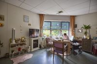 Een bewoonster zit bij het raam aan tafel in haar ingerichte woning. Haar rollator staat naast haar stoel.