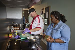 Een man snijdt groenten in de keuken, een vrouw staat naast hem en opent en pot bruine bonen.