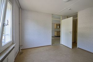 Een lege kamer met beige vloerbedekking, witte muren en een open witte deur die toegang geeft tot een andere ruimte met zichtbare keukenkastjes. Aan de linkerkant bevindt zich een raam.