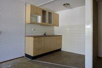 Eenvoudige kitchenette met houten kastjes, betegelde achterwand en houten vloer, in een gedeeltelijk onafgewerkte ruimte.