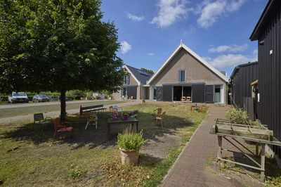 Op de voorgrond een grasveld met een aantal stoelen. op de achtergrond het gebouw met een puntdak en openslaande deuren.