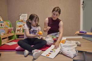 Een meisje en een vrouw zitten op een mat in een ruimte met kasten met kinderboeken. Ze kijken samen naar een boek met plaatjes van fruit.