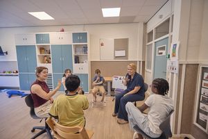 Vier kinderen en twee vrouwen zitten op stoelen in een kring in een kleurrijk klaslokaal.