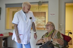 Een verpleegkundige staat naast een bewoonster in een rolstoel, ze kijken elkaar lachend aan.