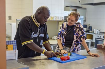 Een man is groente aan het snijden in de keuken, een vrouw staat naar hem en kijkt toe.
