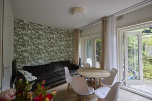 Een woonkamer heeft behang met een groen plantenmotief, voor de muur staat een zwarte bank. Er staat een ronde houten tafel met vijf witte stoelen. De deuren naar de tuin staan open.