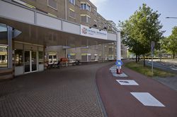 Ingang van een woonzorgcentrum met een overdekte oprit, een verhard voetpad en een fietspad. Bomen en verkeersborden zijn in de buurt zichtbaar.