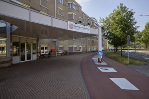 Ingang van een woonzorgcentrum met een overdekte oprit, een verhard voetpad en een fietspad. Bomen en verkeersborden zijn in de buurt zichtbaar.