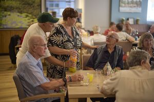 Een groepje ouderen zit aan een gedekte tafel en krijgt drinken aangeboden.