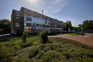 Een modern appartementencomplex met grote ramen onder een helderblauwe hemel, omgeven door groene struiken en met een fietspad op de voorgrond.