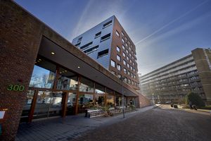 Modern, meerlaags gebouw van baksteen en glas met reflecterende ramen, gelegen op een zonnige dag met een helderblauwe hemel.