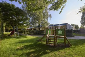 Speeltuin met glijbanen en klimtoestel op gras, omgeven door bomen en struiken. Onder een heldere hemel is op de achtergrond een gebouw zichtbaar.