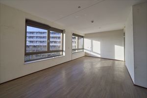 Lichte, lege kamer met een lichte houten vloer, grote ramen aan twee zijden en uitzicht op een woongebouw.