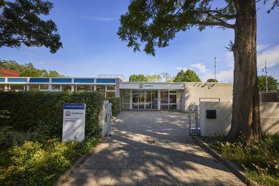 Een modern gebouw met een glazen entree, omgeven door bomen en groen, onder een helderblauwe hemel. Bij de ingang is een bord met de tekst Cordaan de Schuit zichtbaar.
