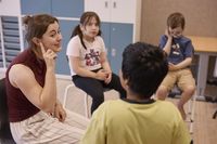 Drie kinderen zitten in een kring in een lokaal samen met een jonge vrouw die naar haar oor wijst.