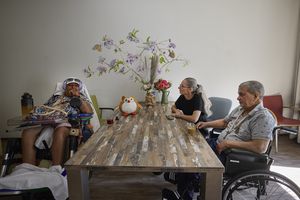 Drie mensen waarvan 2 in een rolstoel zitten aan tafel in de woonkamer. Op tafel staan bloemen.