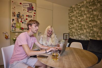 Een begeleider zit aan tafel met een bewoner, hij laat haar iets zien op de laptop.